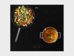 Zwarte inductiekookplaat van ETNA met digitale bediening, waarop een wokpan met groenten en een pan met soep of stoofgerecht staan te koken