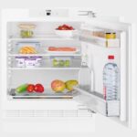 Open koelkast gevuld met diverse voedingsmiddelen, waaronder groenten, fruit, yoghurt, zalm, eieren, een potje mosterd, een fles Vittel-water en een glazen fles.