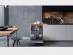 Moderne open keuken met ingebouwde, openstaande vaatwasser vol servies, strakke witte onderkasten, houten werkblad en betongrijze achterwand.