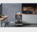 Moderne open keuken met ingebouwde, openstaande vaatwasser vol servies, strakke witte onderkasten, houten werkblad en betongrijze achterwand.